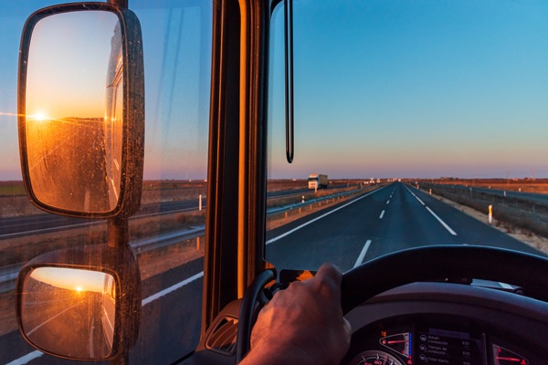 A semi-truck cab interior shows a non-identifiable driver heading down a Texas highway. The dashboard, mirrors, and road ahead are visible, with a green “Entering Texas” sign along the roadside and open fields under a bright sky.