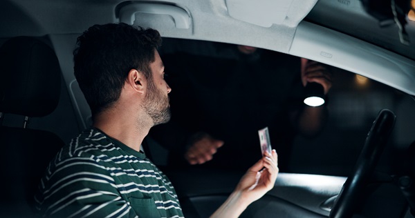 A driver hands a license to a police officer shining a flashlight during a nighttime DWI traffic stop.
