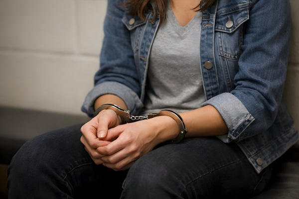 A woman in a denim jacket and grey t-shirt sitting with her hands handcuffed in front of her.