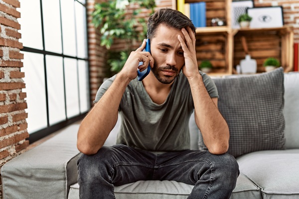 A distressed man sitting on a couch, holding his head in one hand while talking on a smartphone.