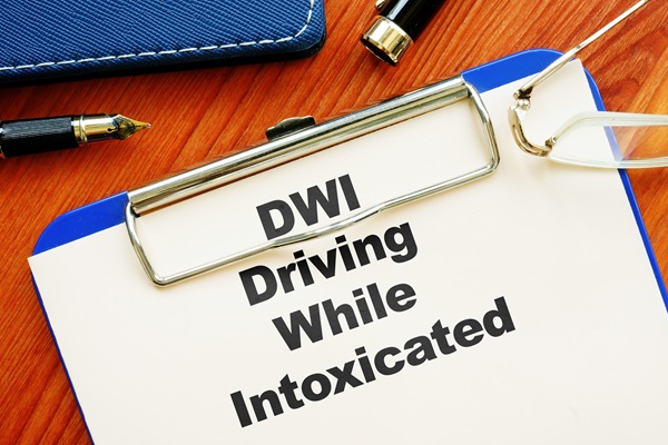 A blue clipboard on a wooden surface holding a document with the text "DWI Driving While Intoxicated" in bold letters, surrounded by a fountain pen and eyeglasses.