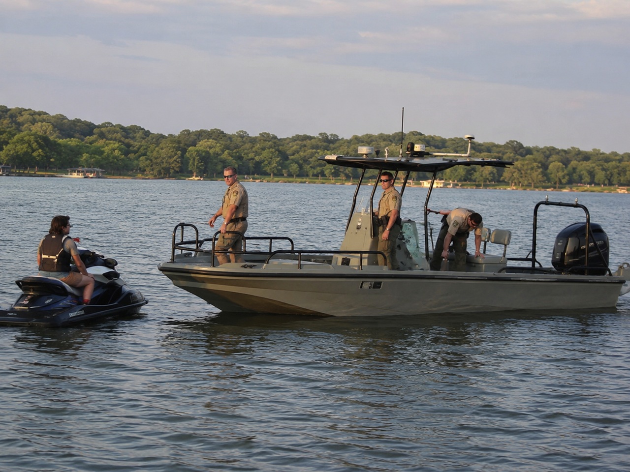 Three law enforcement officers aboard a patrol boat pull up alongside a person riding a jet ski on a large lake lined with green trees.